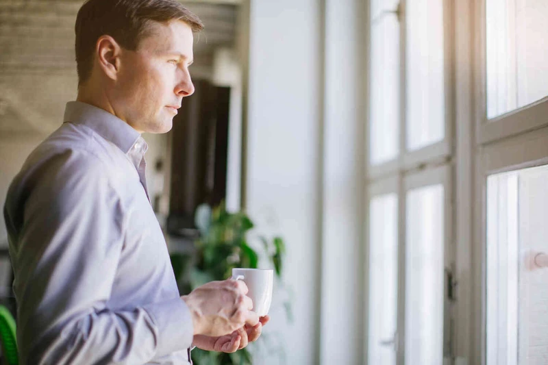 Mann steht mit Becher vor dem Fenster und guckt nach drau&szlig;en